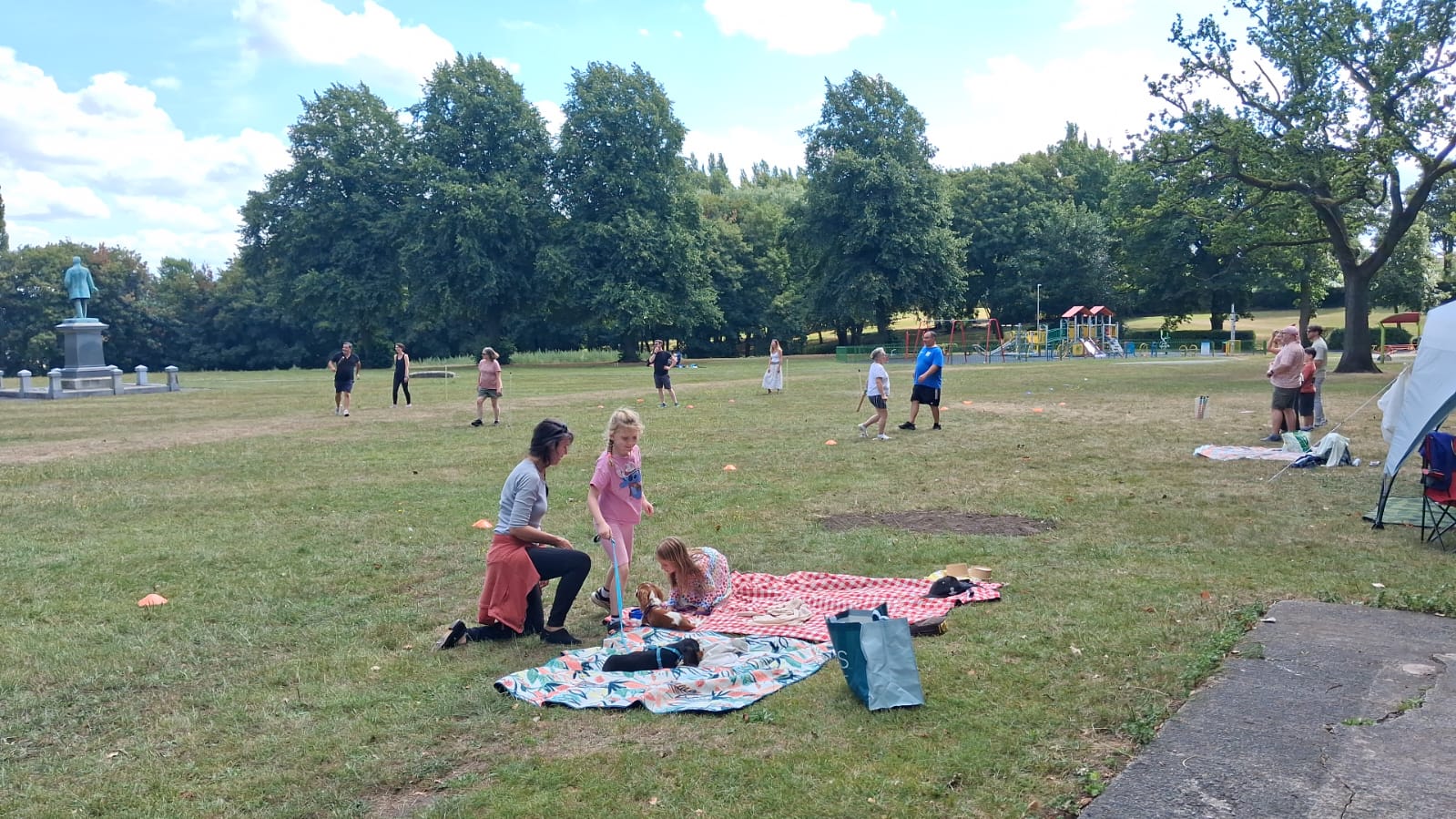 Families relaxing on picnic blankets on the lawn at Verdin Park, with the play area and statue in the distance